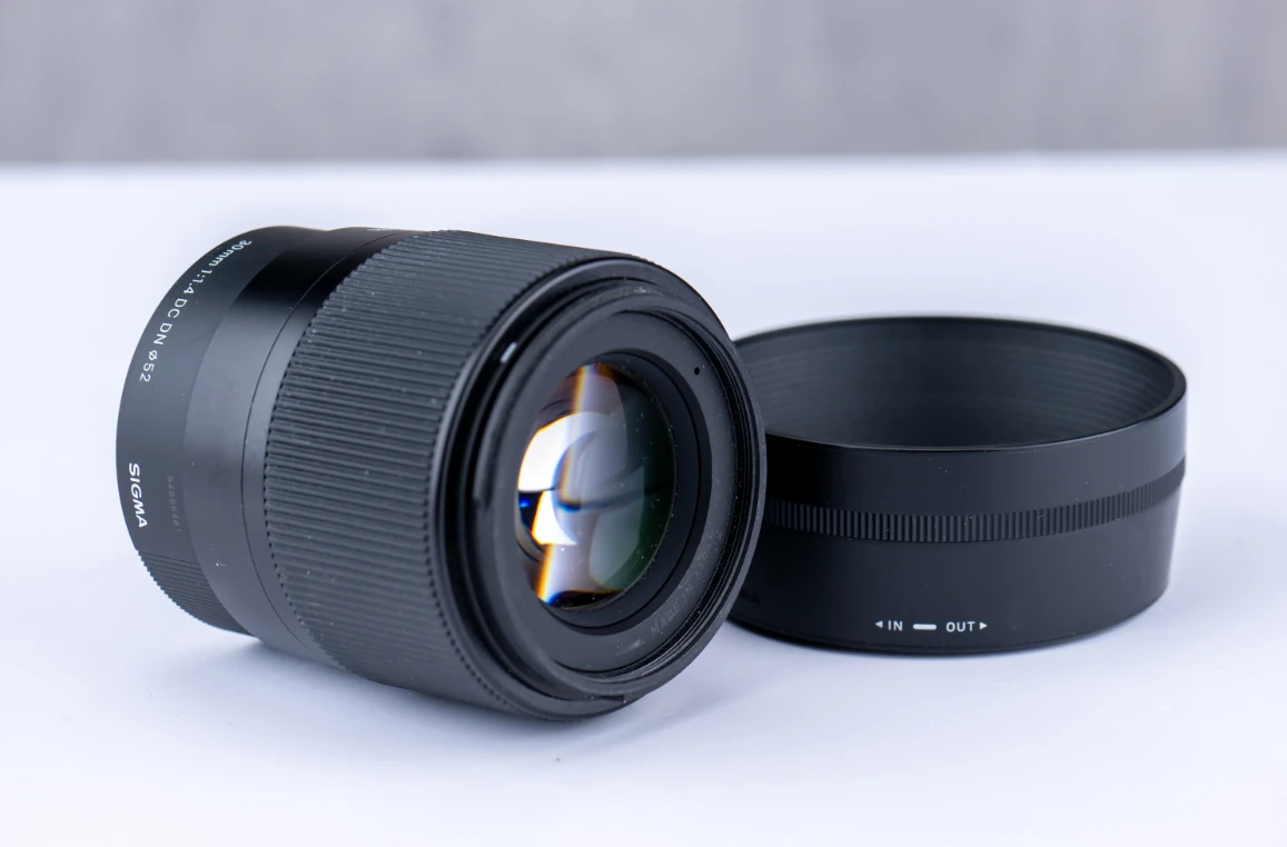 Об'єктив Sigma 30mm f/1.4 DC DN лежить на білій поверхні поруч із відокремленою блендою, чітко видно передню лінзу з покриттям антивідблиску