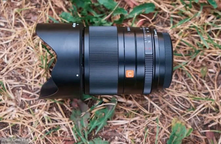 Металевий об'єктив Viltrox AF 33mm f/1.4 з надітою блендою лежить на траві, видно механічне кільце діафрагми зі значеннями від 1.4 до 16 і позицією A