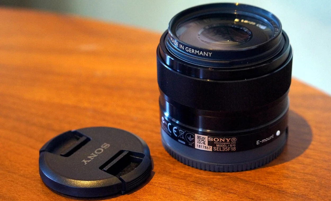 Об'єктив Sony E 35mm f/1.8 OSS стоїть вертикально на лакованому столі поруч із фірмовою передньою захисною кришкою з логотипом Sony