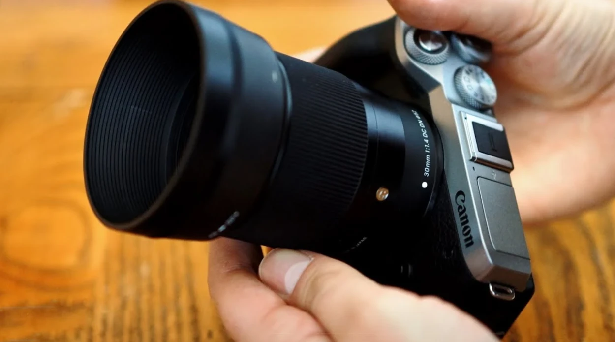 Об'єктив Sigma 30mm f/1.4 DC DN з блендою у руках оператора, приєднаний до компактної системної камери з видимою шкалою 52 мм