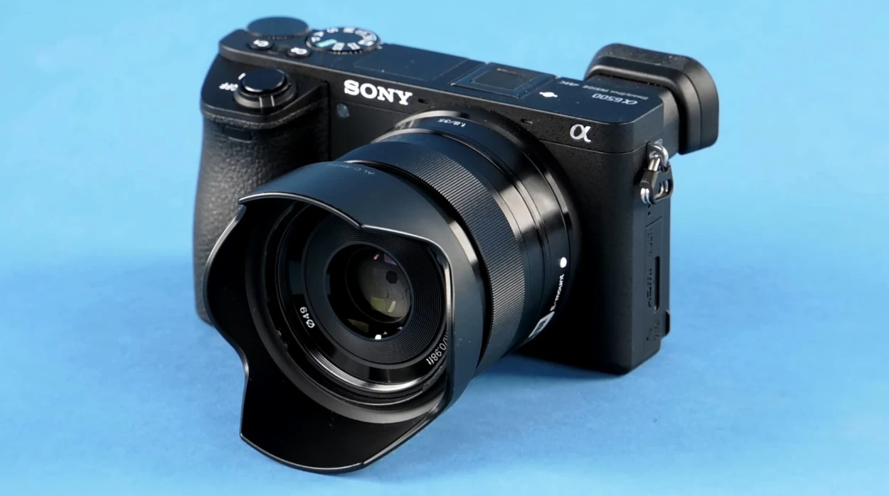 Компактний чорний об'єктив Sony E 35mm f/1.8 OSS з пелюстковою блендою ALC-SH112 на камері Sony alpha 6500 з маркуванням SteadyShot INSIDE на синьому фоні